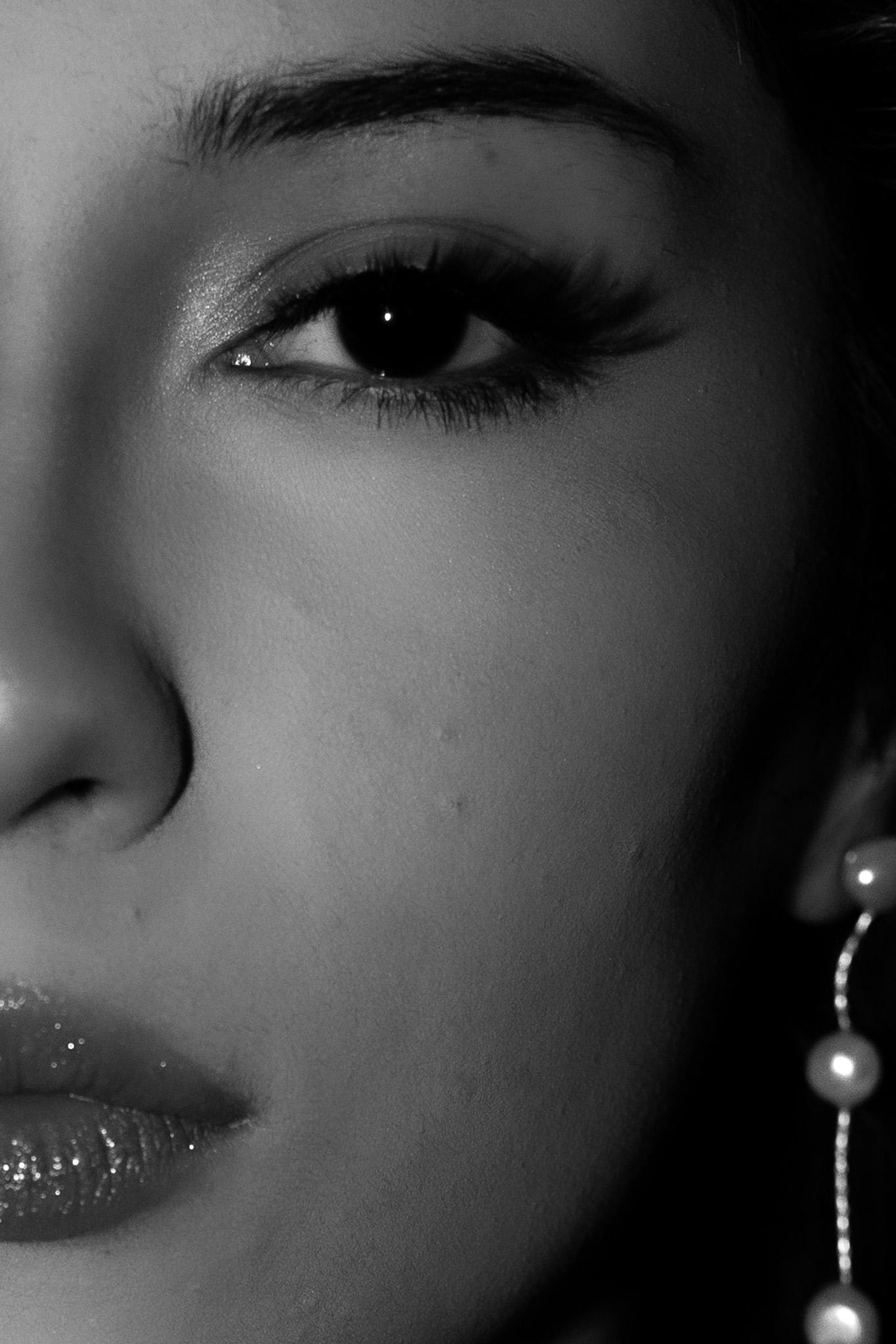 Black-and-white macro beauty portrait of a woman's eye, cheek and glossed lips with a pearl drop earring fading into shadow