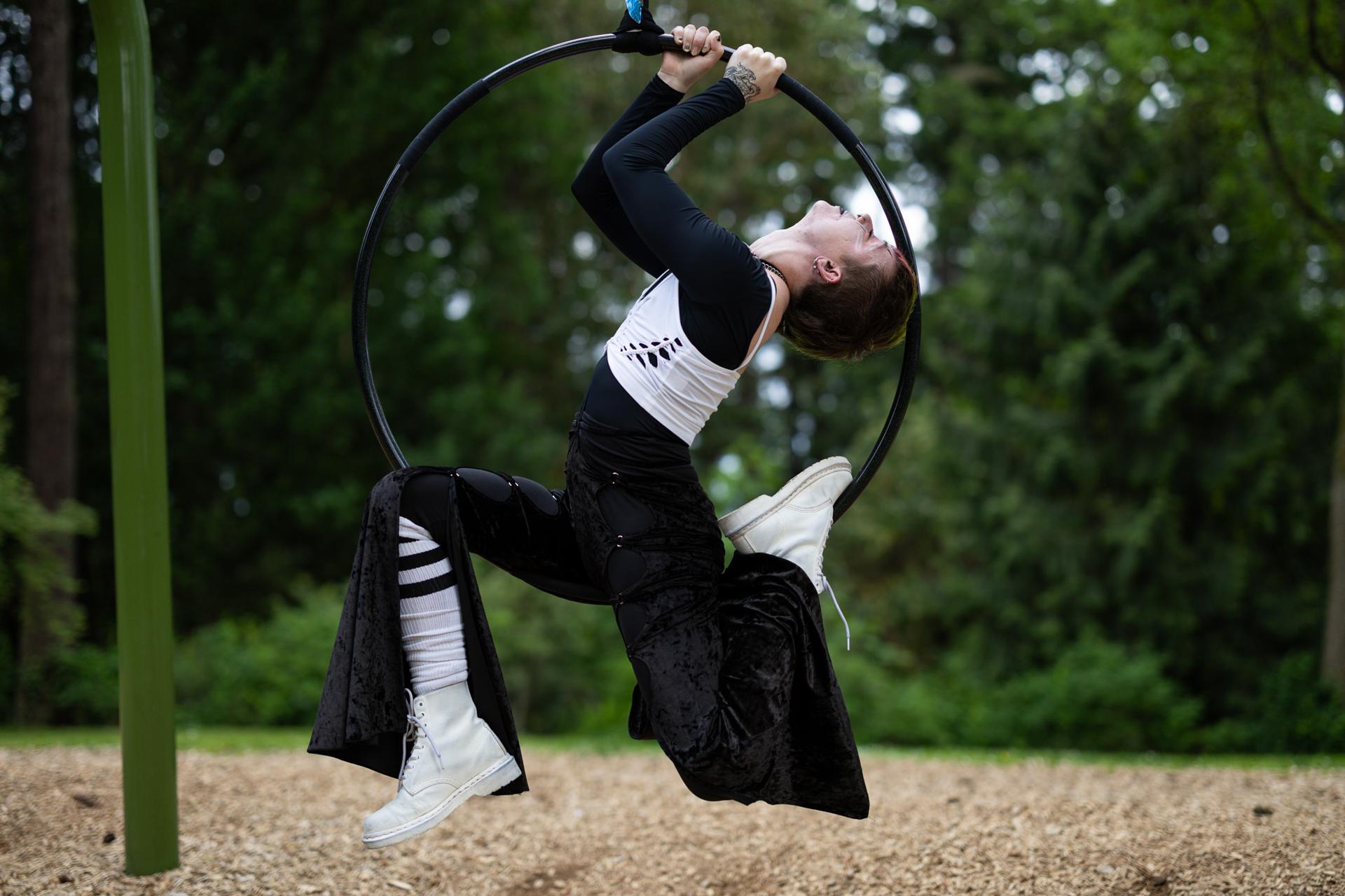 An aerial performer in black velvet and white boots arches backward inside a suspended hoop in a forested park