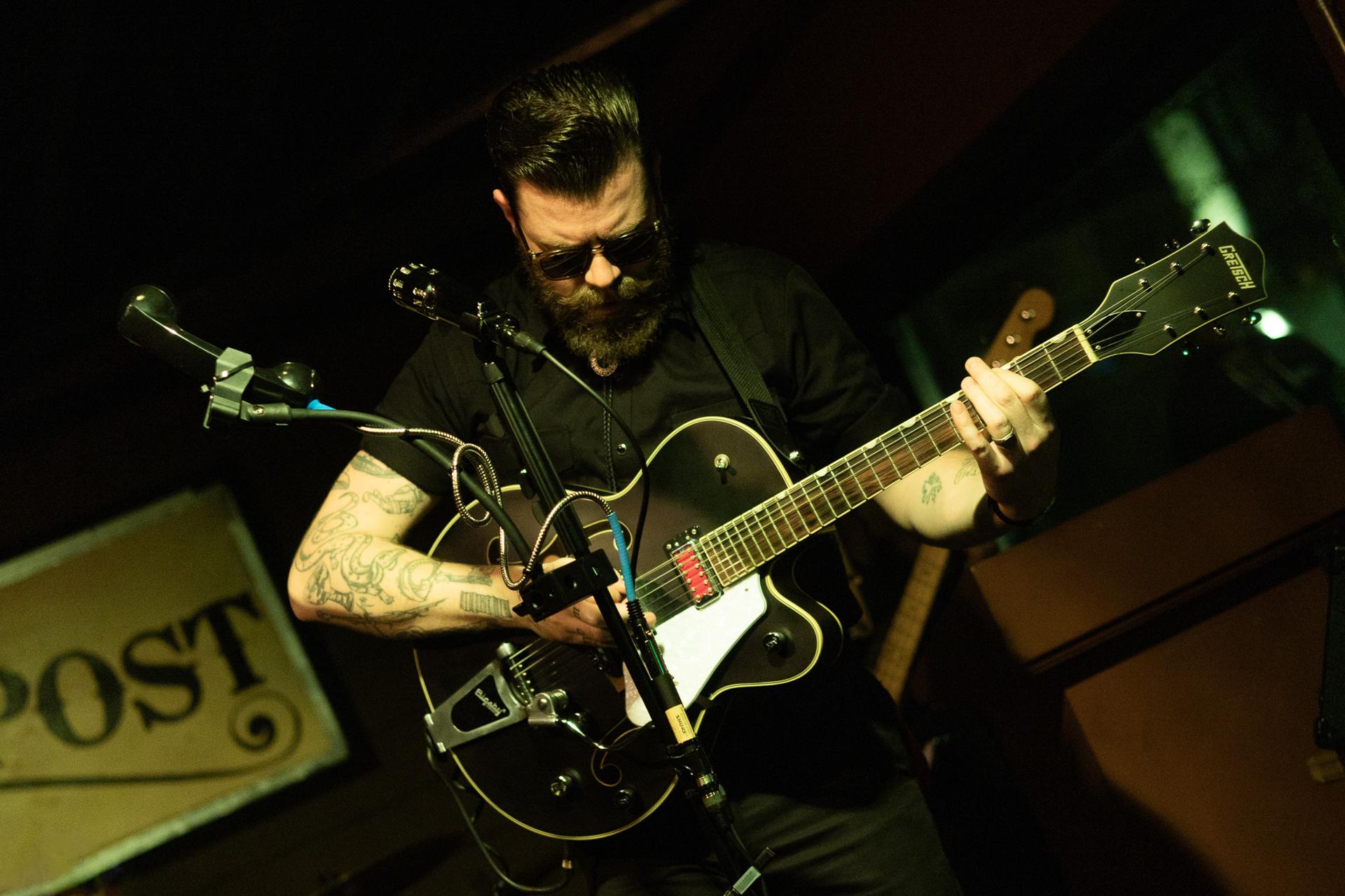 A bearded man in sunglasses and a black shirt playing a black Gretsch guitar on a dimly lit stage beside a vintage POST sign