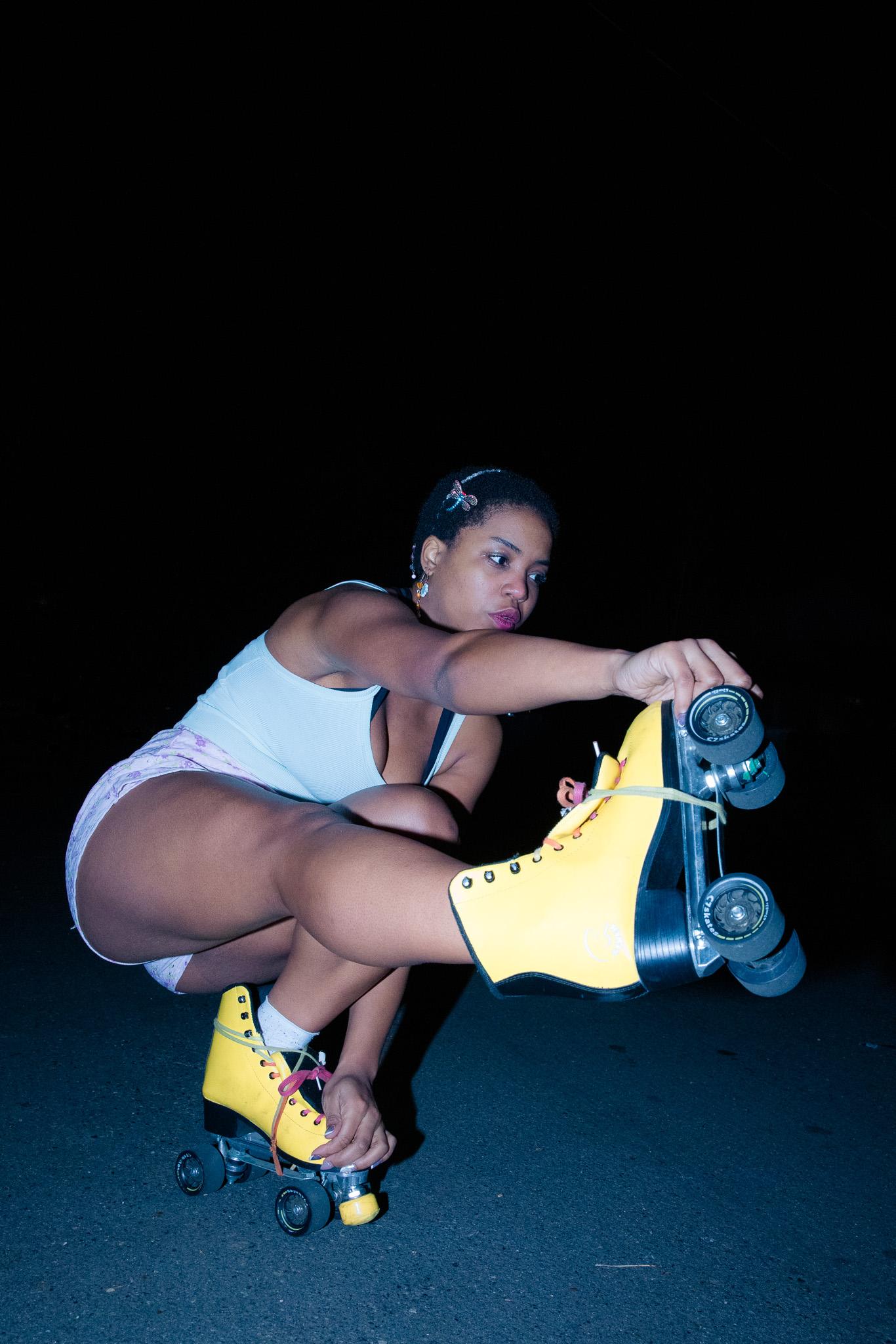 Woman in a white tank top and shorts crouched low on dark pavement at night lacing a pair of vivid yellow roller skates, looking back over her arm toward the camera