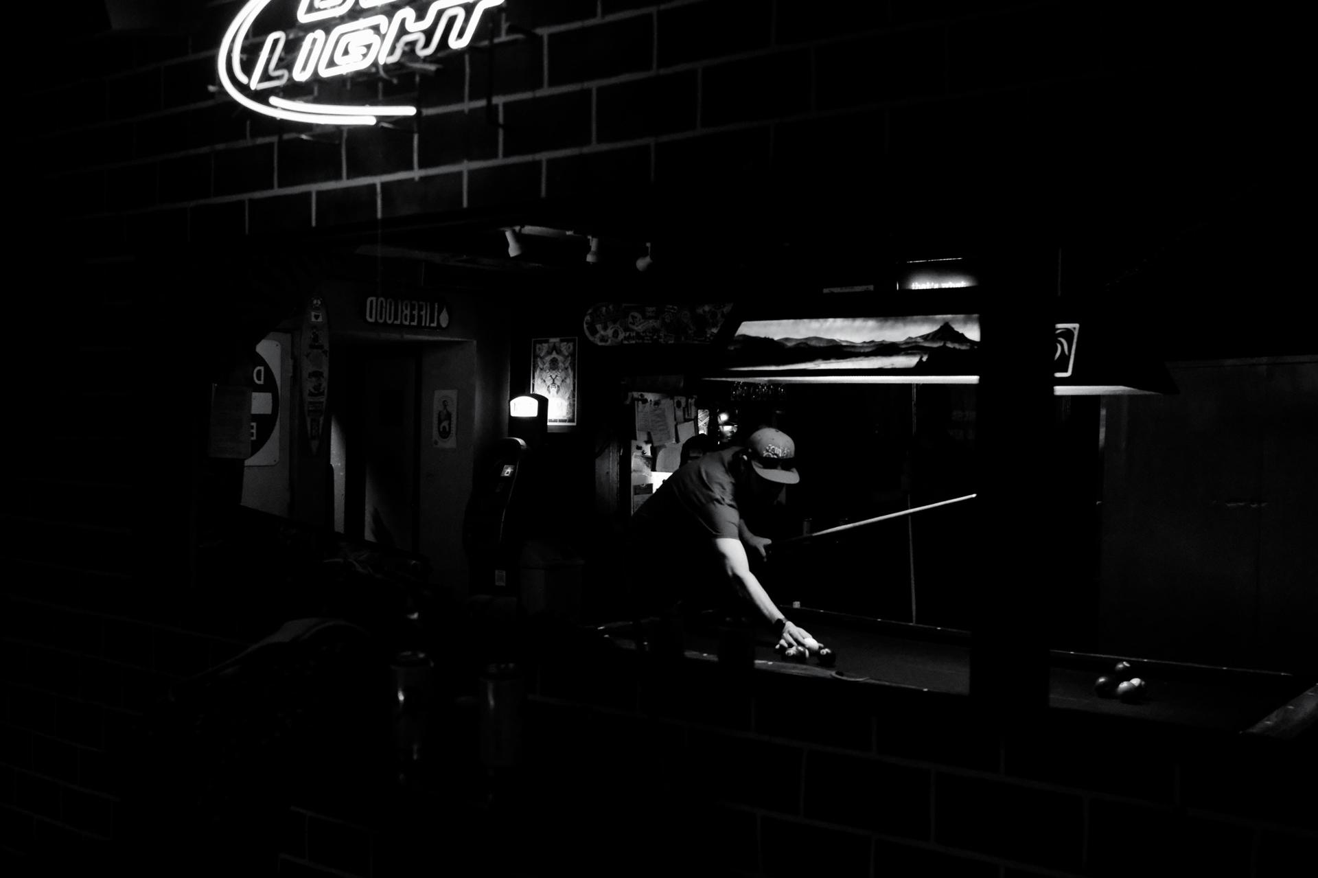 A man in a cap leans across a pool table to take a shot in a dark dive bar lit by a Bud Light neon sign