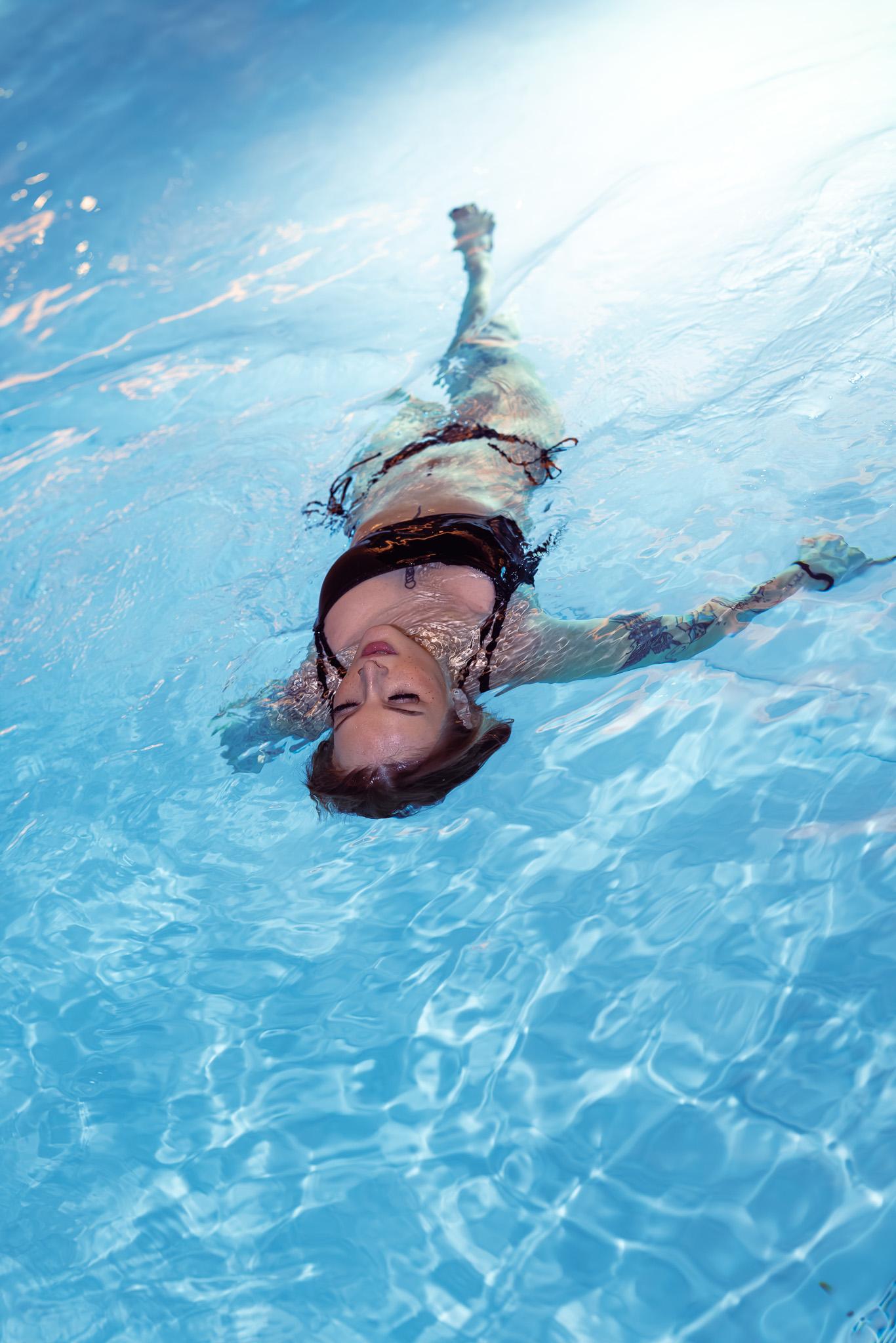 A woman floats face-up in bright turquoise pool water, her body trailing diagonally with caustic ripple light distorting her face and a tattooed arm extended