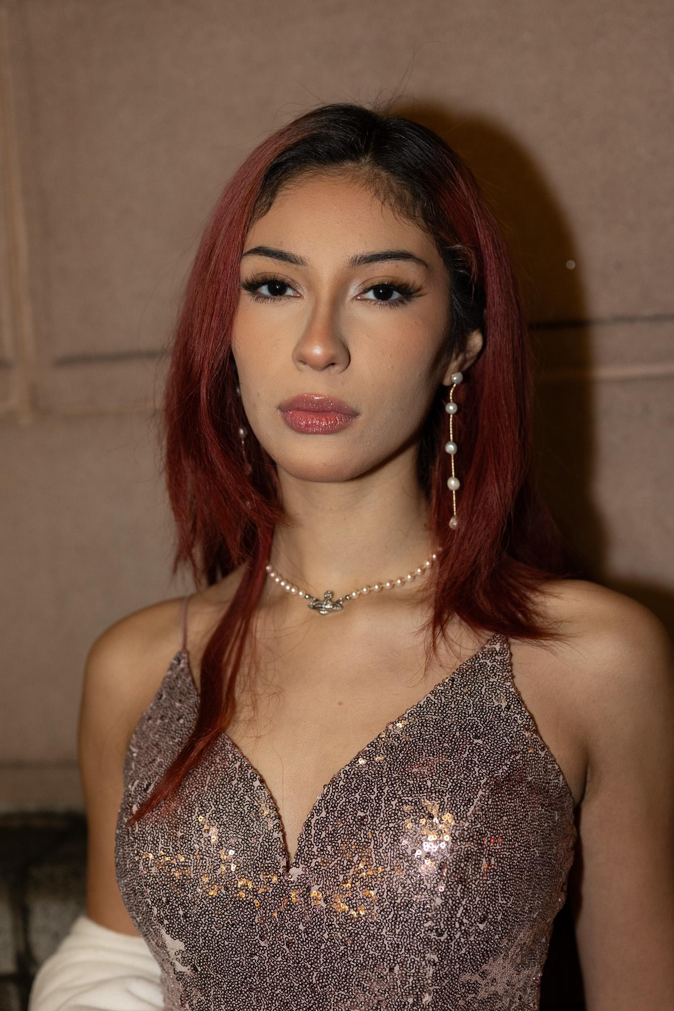 Red-haired model in a rose sequined slip dress with a pearl choker and pearl drop earrings, gazing straight into the camera against a warm sandstone wall