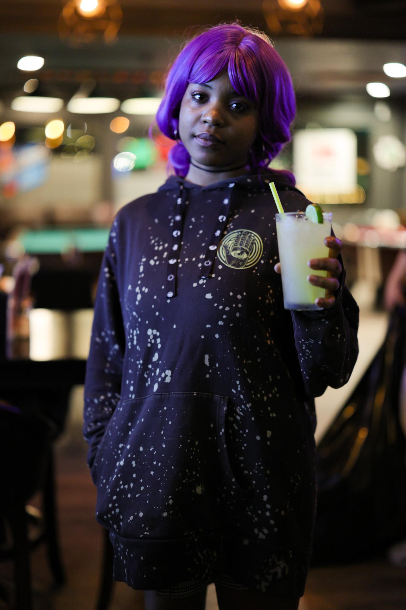Woman in a vivid purple wig and dark paint-splattered hoodie holding a pale cocktail with a lime, standing in a warmly lit bar with glowing bokeh behind her