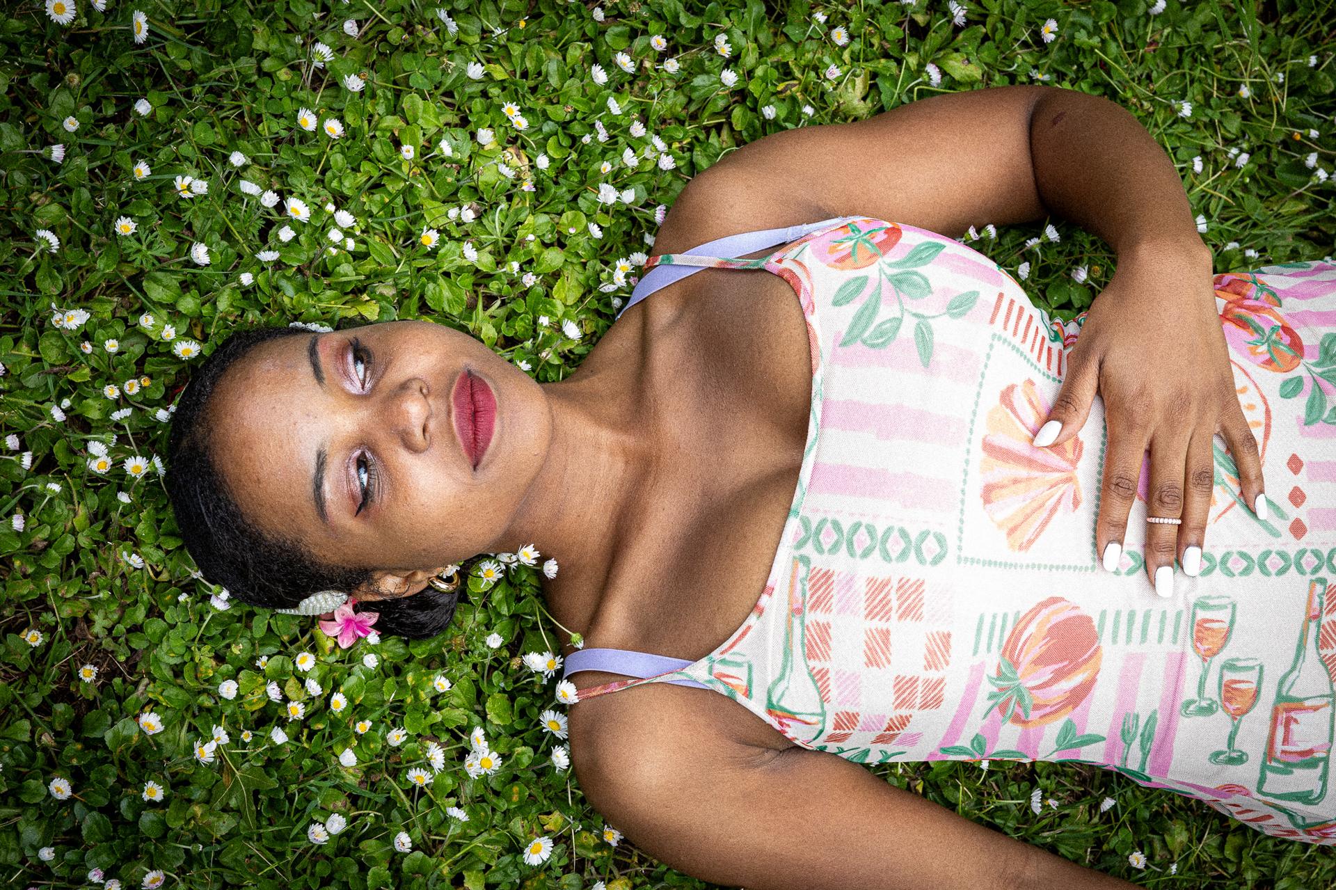 Overhead portrait of a woman lying in green clover and white daisies wearing a pink-printed tank top, gazing upward with a flower beside her face