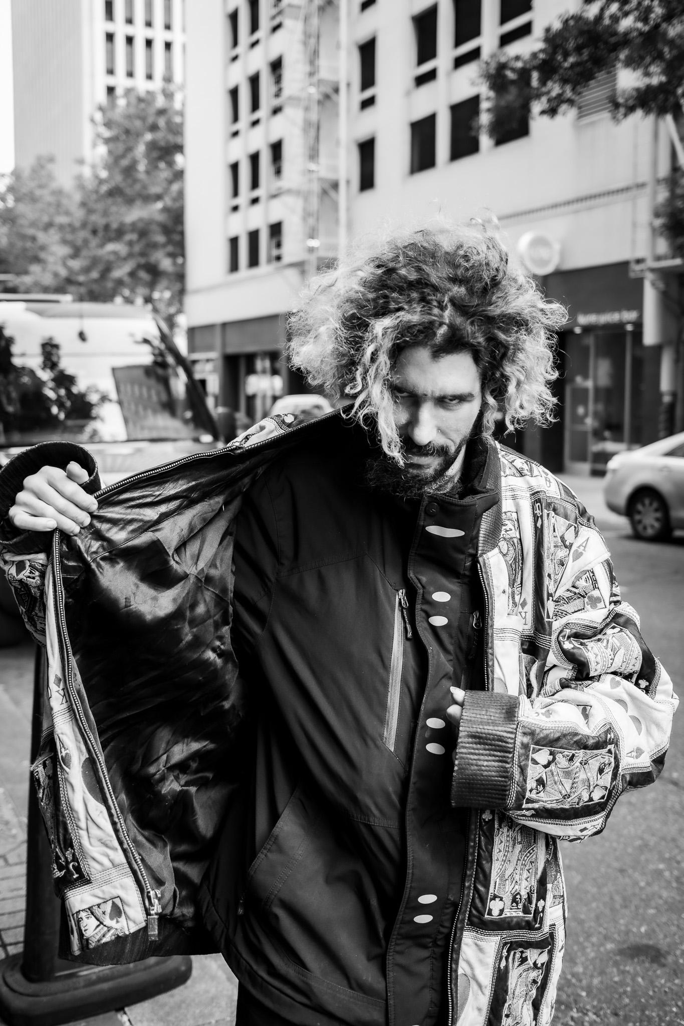 Black-and-white street portrait of a man with voluminous curly hair opening a patterned black bomber jacket on a city sidewalk
