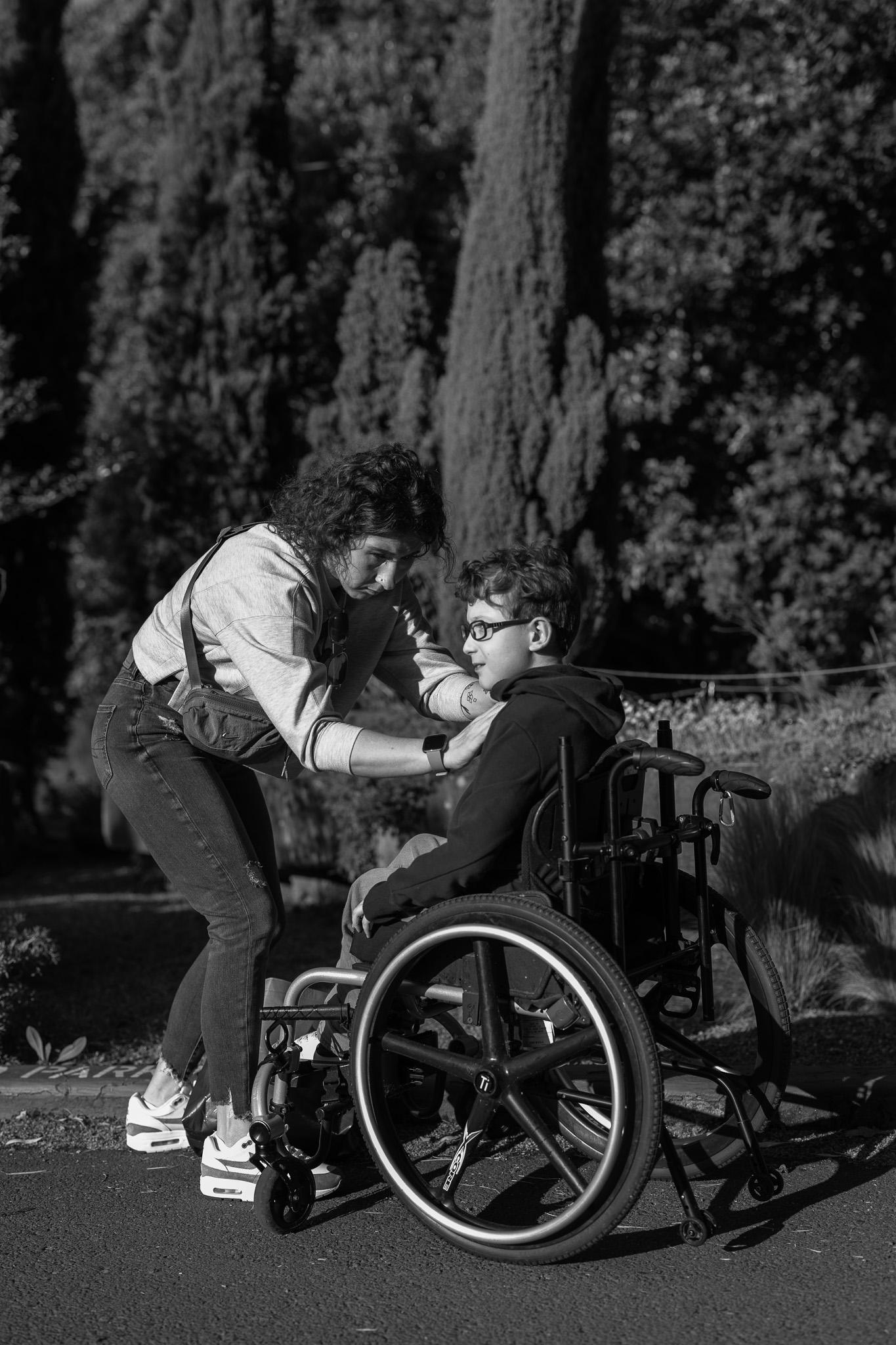 Black and white photo of a young woman leaning down to connect with a smiling boy in a wheelchair on a tree-lined park path