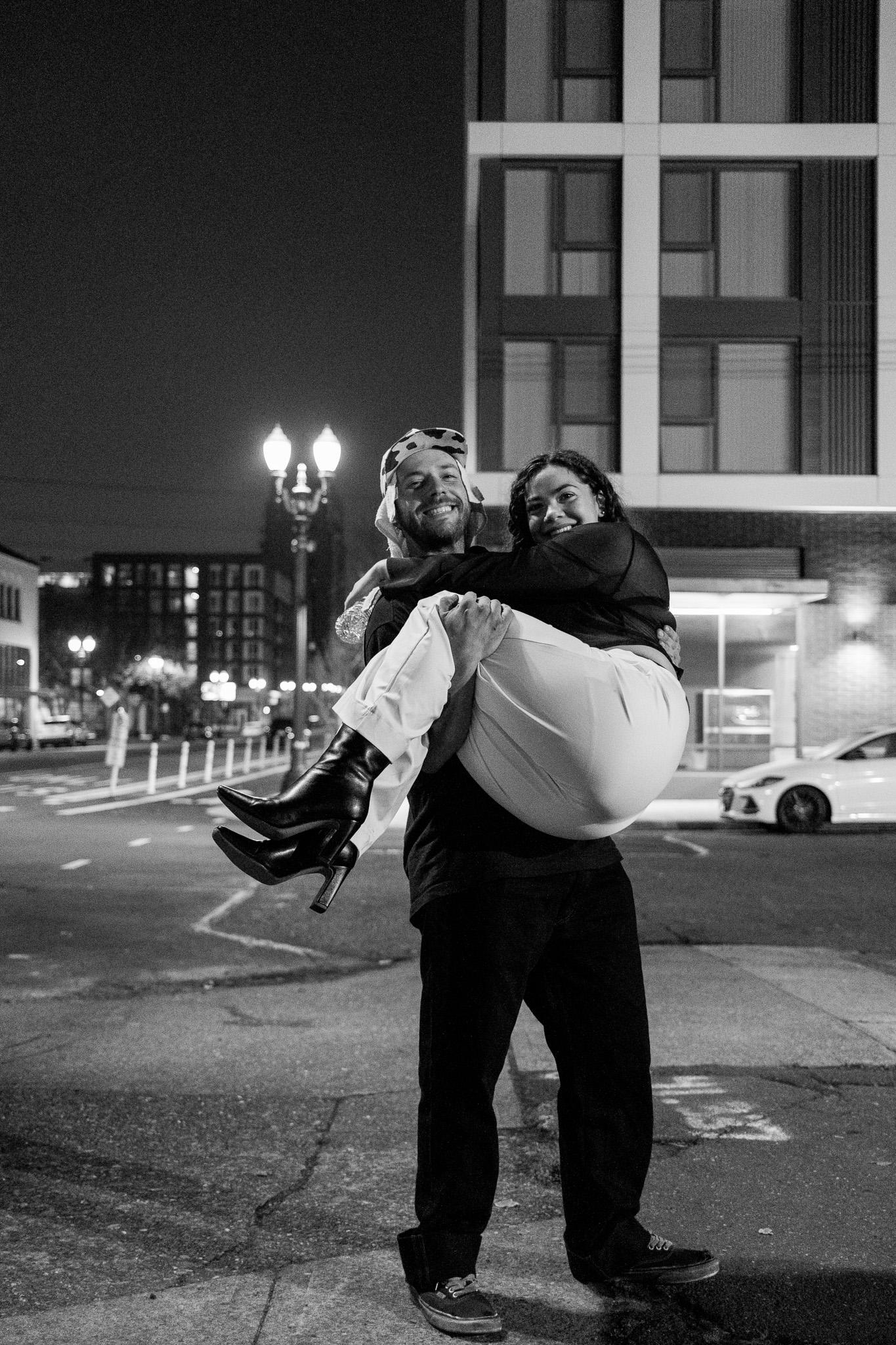 Man in dark clothes carrying a laughing woman in white pants and black heeled boots across his arms on a city street at night, a lit high-rise and street lamp behind them