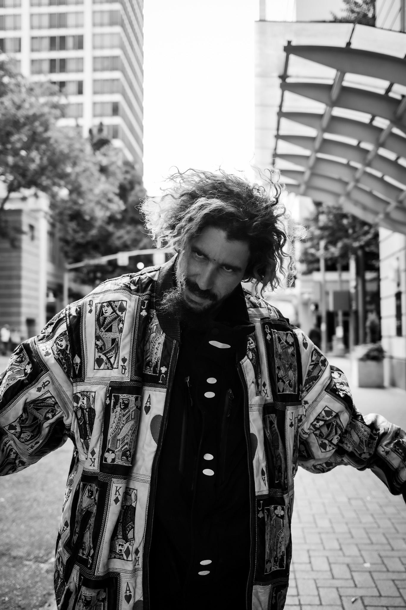 Man with windblown hair in a playing-card print jacket leaning toward the camera on a sunlit downtown plaza
