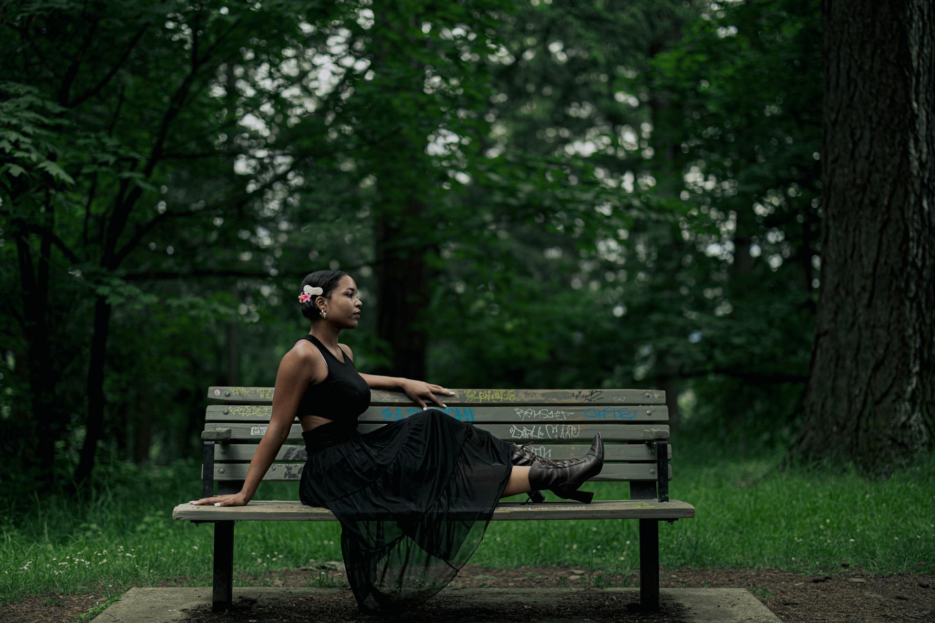 A woman in a black top and sheer black skirt with cowboy boots reclines in profile on a weathered park bench surrounded by deep green forest